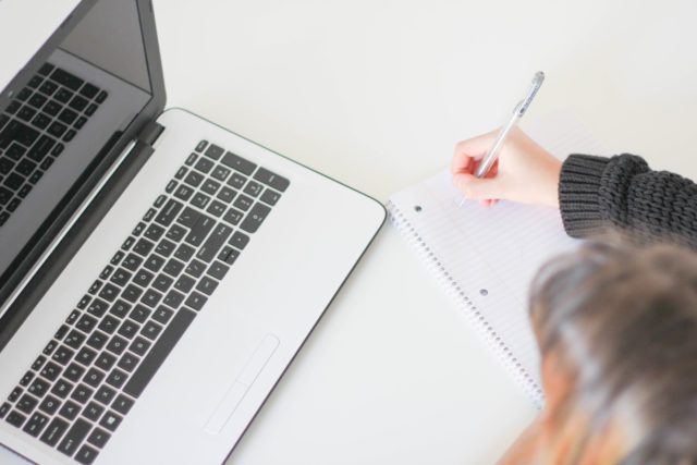 A woman writing on a notebook next to a laptop.